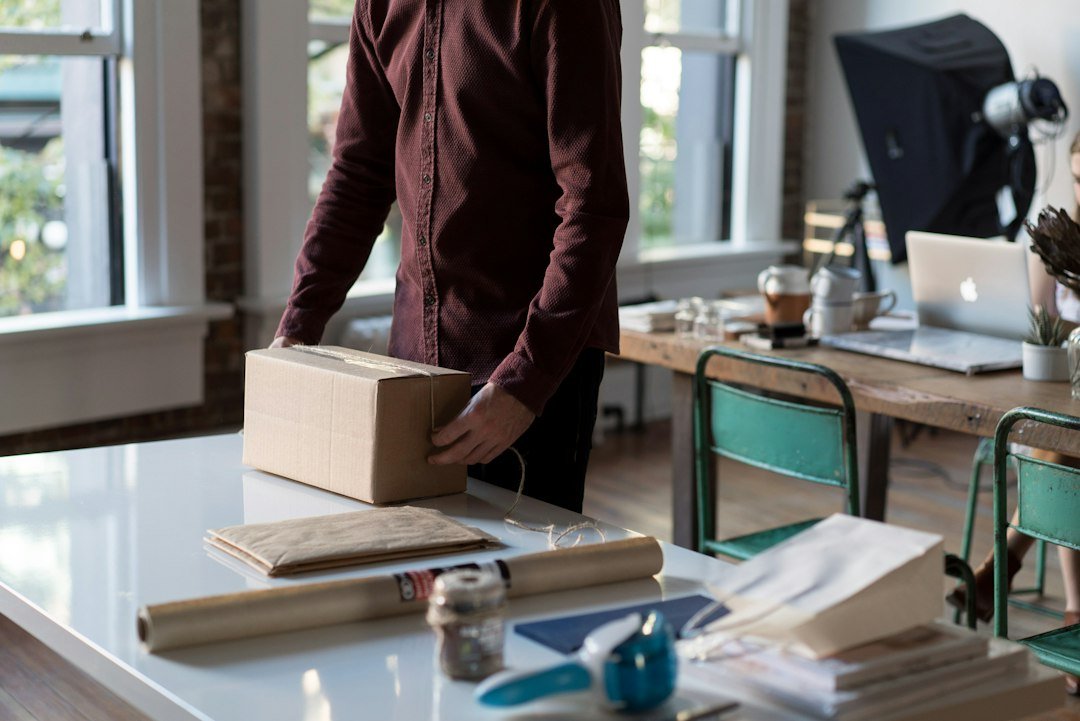 Person holding a cardboard parcel on a packing table, with a laptop behind it